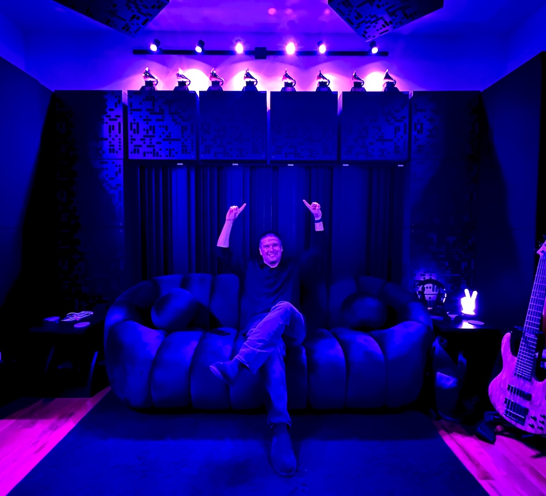 Adam Ayan sitting at the rear of his new mastering studio, pointing to the eight Grammy and Latin Grammy Awards atop the back wall.